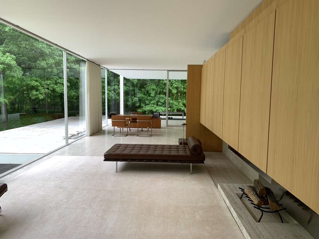 A minimalist, modernist interior space featuring floor-to-ceiling glass walls that bring the surrounding forest into the living area. The room showcases a Barcelona-style leather daybed, light wood wall paneling, and a seamless flow between indoor and outdoor spaces. The natural light, clean lines, and careful material selection exemplify mid-century modern design principles with attention to architectural details.